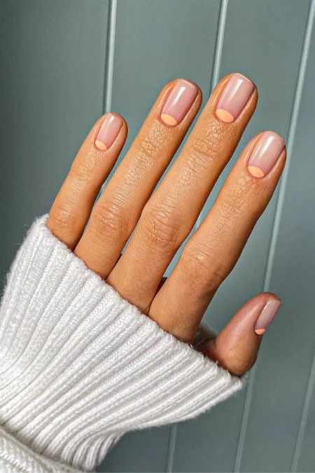 Close-up of well-manicured nails with a short nail design with nude polish on a hand wearing a white knitted sweater, against a muted green background.