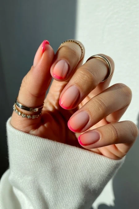 Close-up of a hand with manicured nails featuring a natural base and bright pink tips, adorned with multiple rings, against a neutral background.