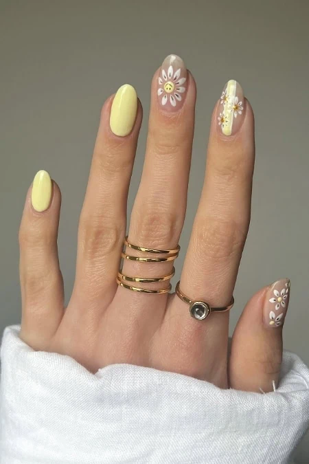 Close-up of a hand with pastel yellow and floral nail art, featuring daisy designs and gold rings, showcasing trendy spring nail fashion and an example of short nail designs.