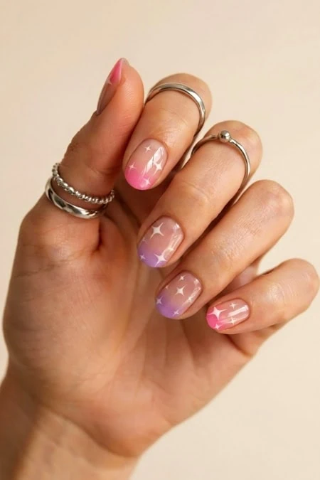 Colorful star-themed nail art with pastel pink, purple, and white shades on manicure, accessorized with silver rings, perfect for beauty and fashion enthusiasts.