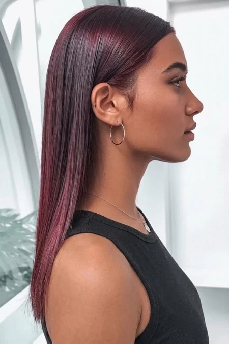 Elegant woman with sleek, straight burgundy cherry cola hair color, wearing hoop earrings and a black sleeveless top, standing indoors with natural light, showcasing modern beauty and hair styling.