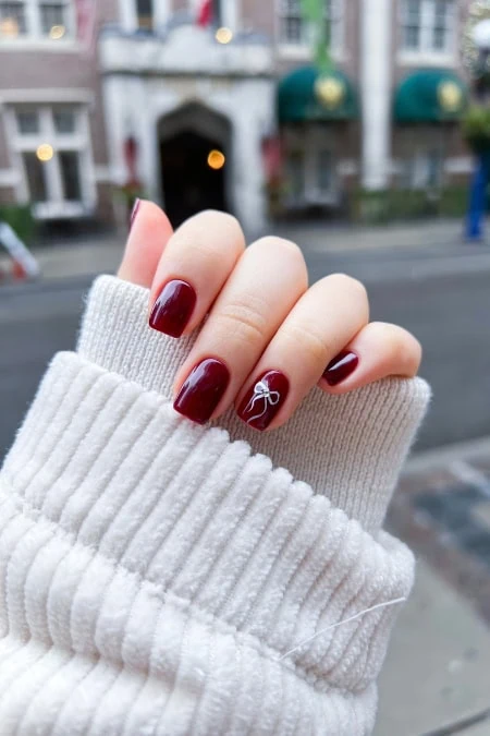 Close-up of a woman's hand with manicured nails painted deep red, featuring a white bow design on the ring finger, against an urban street background showcasing example of short nail designs.