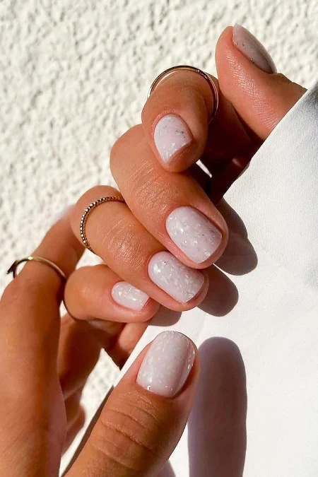 Close-up of a hand with well-manicured nails painted in a soft pink shade, adorned with delicate rings, against a textured white background.