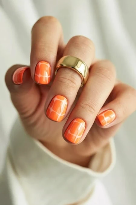 Close-up of a woman's hand with orange geometric patterned nail polish and a gold ring, showcasing trendy nail art and elegant jewelry.