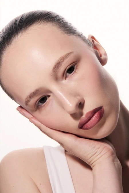 Close-up of a young woman with flawless skin, natural fall makeup look, and a serene expression, resting her face on her hand against a white background.