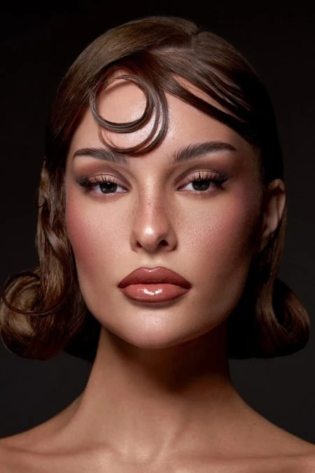 Close-up of a woman with vintage-inspired fall makeup and hairstyle, featuring glossy lips, defined eyebrows, and a styled curl on her forehead, against a dark background.