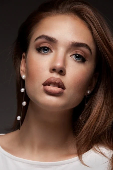 Close-up portrait of a young woman with flawless skin, brown hair, and makeup emphasizing her eyes and lips, suitable for beauty and fall makeup.