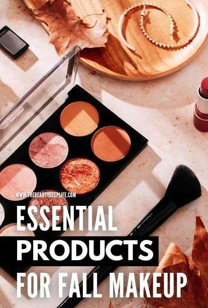 Fall makeup essentials including eyeshadow palette, blush, and brushes arranged on a marble surface with autumn leaves and jewelry for seasonal beauty look.