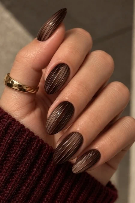 Close-up of a hand with manicured nails painted in dark brown with fine vertical stripes, wearing a gold ring and a maroon sweater sleeve.