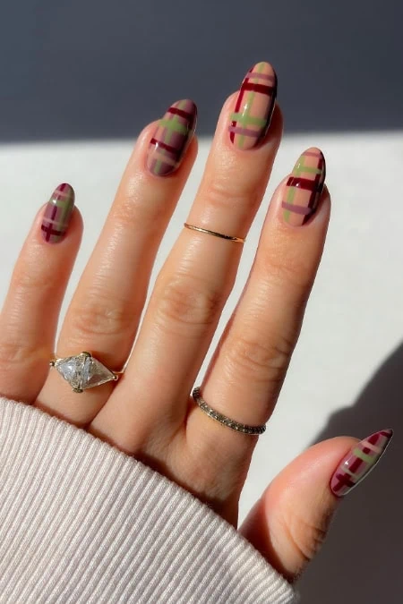 Fashionable nails with plaid pattern in red, green, and beige, adorned with rings, held against a neutral background for a stylish, modern look.