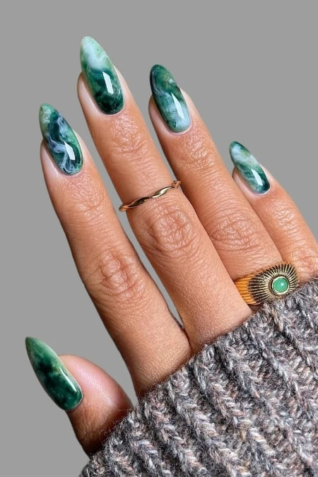 Close-up of a hand with green marble-patterned acrylic nails, wearing a gold ring with a turquoise stone and a textured gray sweater.