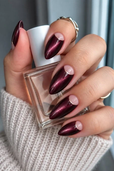 Close-up of a woman's hand with long, almond-shaped, metallic burgundy nails holding a clear nail polish bottle.