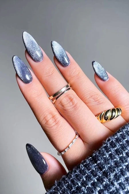 Close-up of a hand with long, almond-shaped nails painted in a silver glitter gradient, adorned with multiple gold and silver rings, against a neutral background.