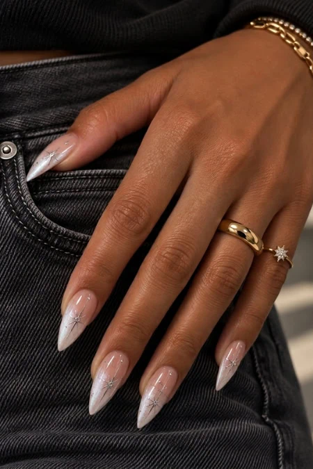 Close-up of a woman's hand with long, elegant acrylic nails featuring a clear base and white tips, adorned with gold rings and a delicate bracelet, against black denim jeans.