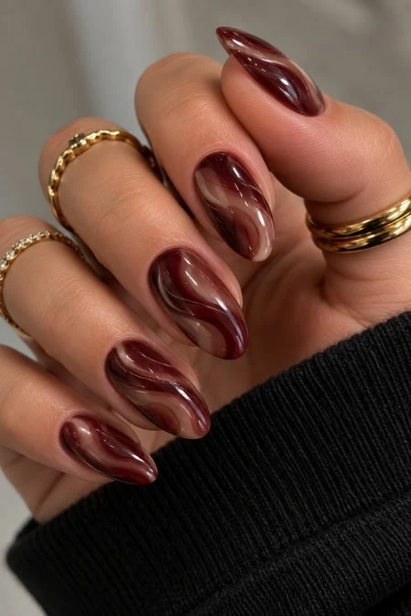 Close-up of a hand with maroon and white marbled acrylic nails, adorned with gold rings, against a black sleeve background.