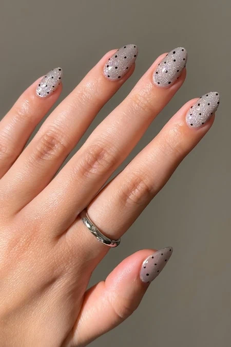 Close-up of a hand with manicured nails painted in light gray with black polka dots, featuring a silver ring on the ring finger, against a plain background.