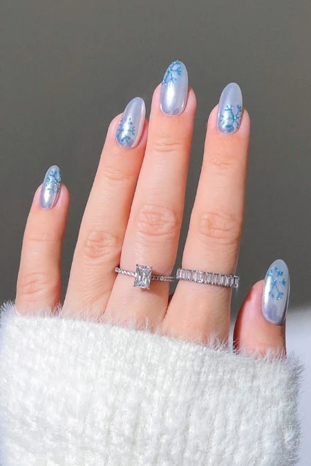 Elegant winter nail art with icy blue and silver snowflake designs on manicured nails, complemented by a silver ring with a large gemstone.