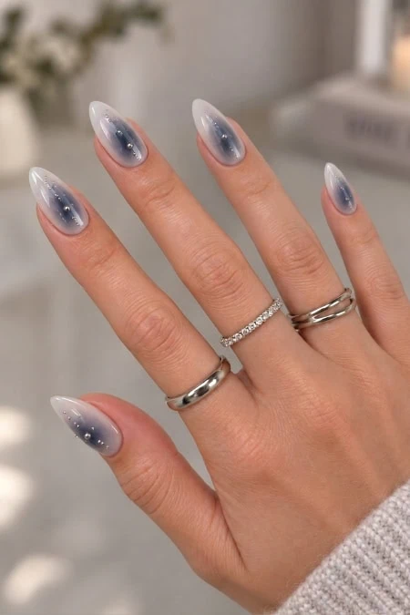 Elegant hand with long, almond-shaped nails featuring a galaxy-inspired design in shades of blue, gray, and white with tiny star-like accents.