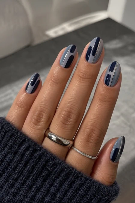 Stylish manicure with navy blue and light gray geometric nail art on well-groomed fingers, featuring rings, against a blurred indoor background.