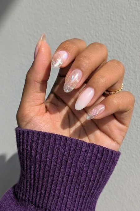 Close-up of a hand with manicured nails featuring glitter and ombre designs, wearing a purple sweater sleeve against a plain background.