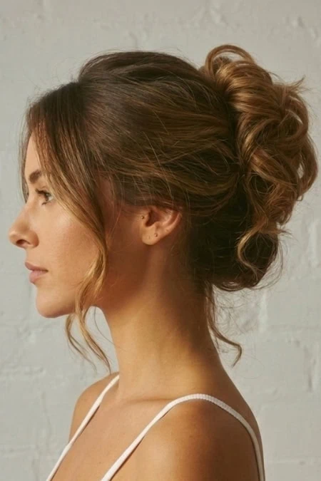 Elegant woman with styled curly updo hairstyle, wearing a white spaghetti strap top, against a neutral background, showcasing beauty and hair styling.