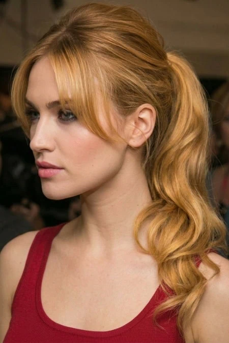 Red-haired woman with styled wavy hair wearing a red sleeveless top, looking to the side in a professional setting.