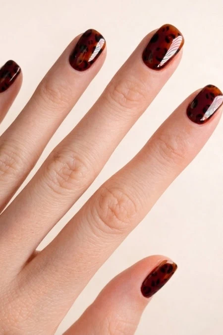 Short winter nails with tortoiseshell nail art on a pale background, close-up manicure showcasing glossy brown-red pattern trend.
