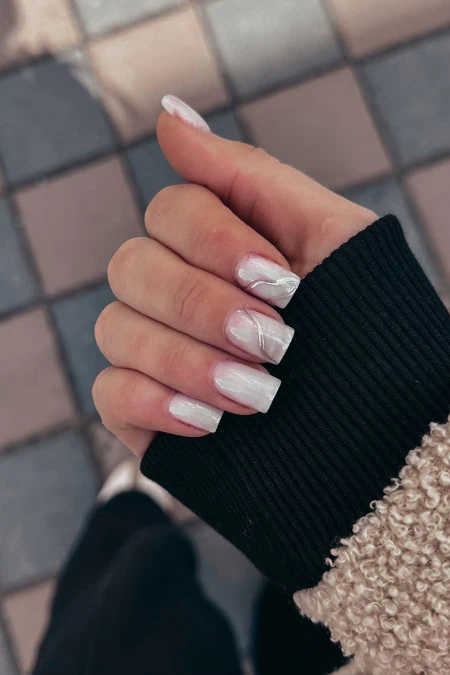 Stylish white marble manicure with square nails and subtle shine, peeking from a black ribbed cuff against a tiled pavement backdrop.