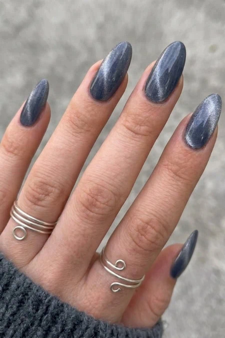 Dark metallic blue-gray nails with long almond shape nail trend and subtle shimmer, worn with minimalist silver rings on a woman's hand.