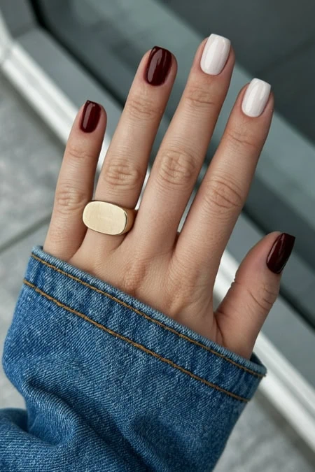 Close-up of a hand with contrasting burgundy and white nails trend for winter, wearing a beige ring and denim jacket sleeve, captured outdoors for a fashion and beauty feature.