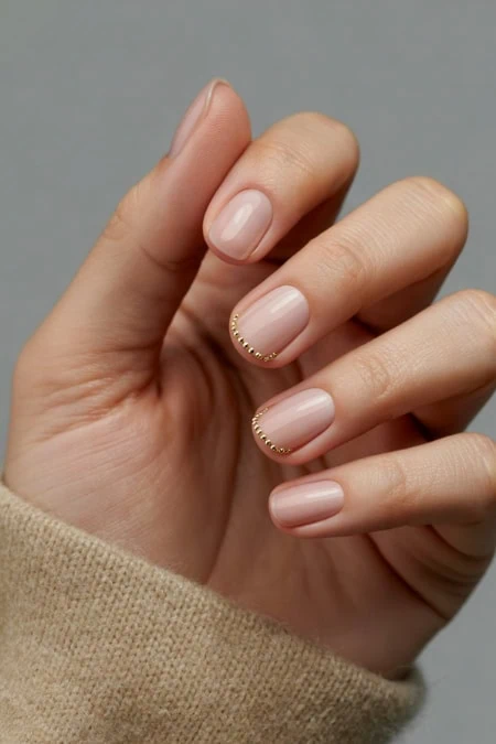 Close-up of a hand with nude pink manicure showcasing winter nail trends and small gold beaded detailing along the cuticle on short, almond-shaped nails.