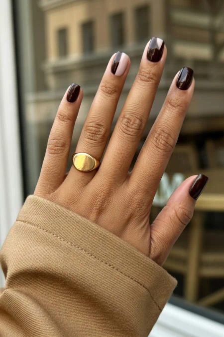 Close-up of a hand with dark brown nail polish wearing a simple gold ring on the ring finger, beige coat sleeve, urban storefront in the background.
