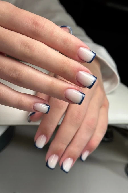 Elegant manicured winter nail trend with a white to light pink gradient and dark blue angular tips, showcasing chic nail art on a model's hands.