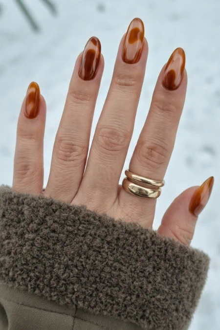 Close-up of a hand with stylish brown tortoiseshell gradient nails wearing two gold rings, wearing a brown cozy coat sleeve against a soft blue background, optimized for Beauty Deep Life styling and秋季.