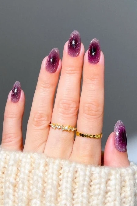 Close-up of hands with short almond nails painted in a gradient purple glitter manicure, sparkly tips and subtle gems, wearing delicate gold rings against a soft gray background for winter nail trends example.