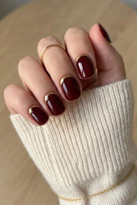 Close-up of a hand with burgundy winter nail trend and a thin gold ring, wearing a cream ribbed knit sweater sleeve.