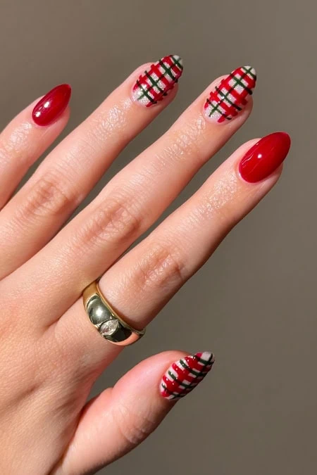 Short elegant winter nails with glossy red tips and plaid patterned accent nails, showcasing seasonal manicure trend on a stylish hand with a gold ring.